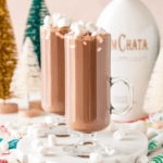Two glass mugs of boozy hot cocoa topped with marshmallows rest on a table, surrounded by colorful miniature Christmas trees, while a bottle labeled "RumChata" hints at the delightful blend.