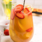 A glass pitcher filled with a refreshing Easy Sangria Mimosas recipe—orange juice, sliced strawberries, and other fruits—graces the marble countertop. A black spoon is submerged in the vibrant drink, inviting you to indulge.