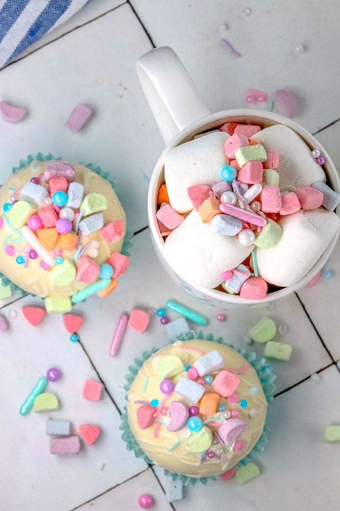 white chocolate hot chocolate bomb in a blue cupcake wrapper with lucky charms marshmallows and sprinkles on top next to a mug with cocoa, sprinkles, and marshmallows in it