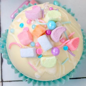 white chocolate hot chocolate bomb in a blue cupcake wrapper with lucky charms marshmallows and sprinkles on top next to a mug