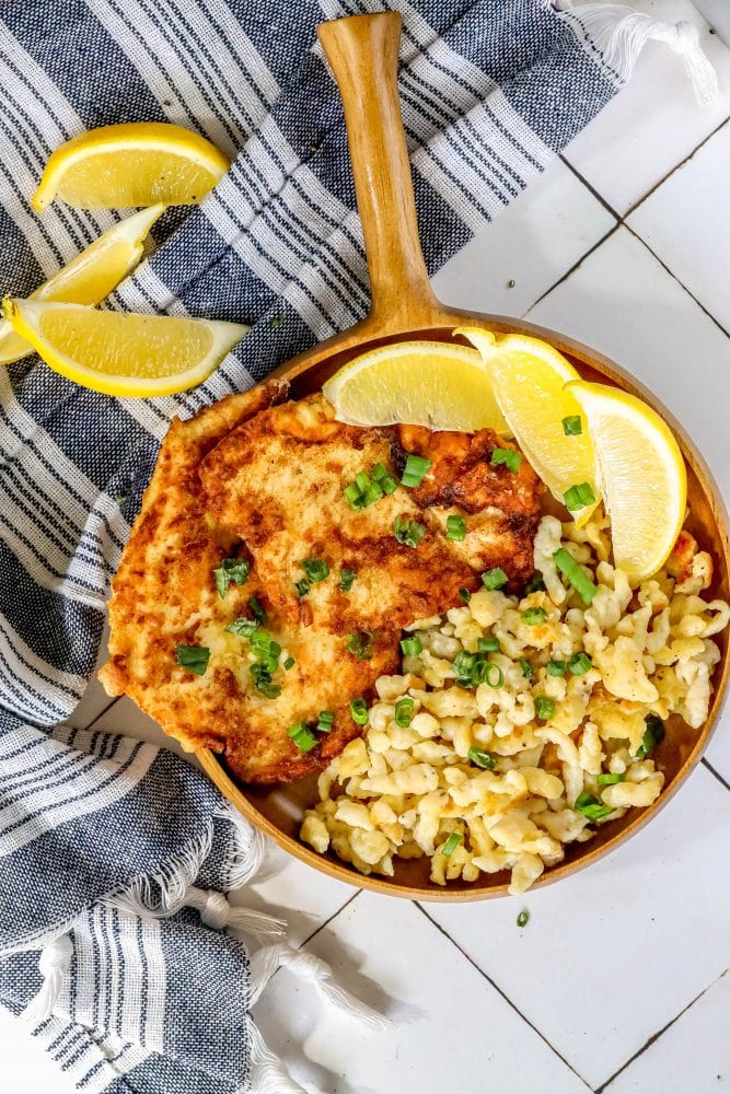 picture of breaded chicken schnitzel in a wooden bowl with green onions on top and lemon slices and spaetzle next to it