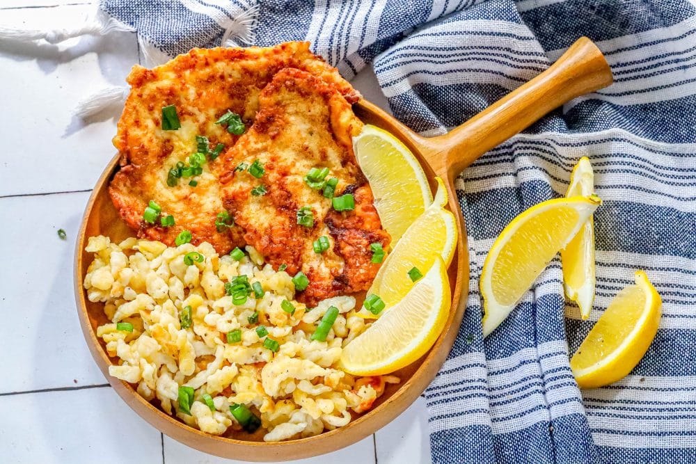 picture of breaded chicken schnitzel in a wooden bowl with green onions on top and lemon slices and spaetzle next to it