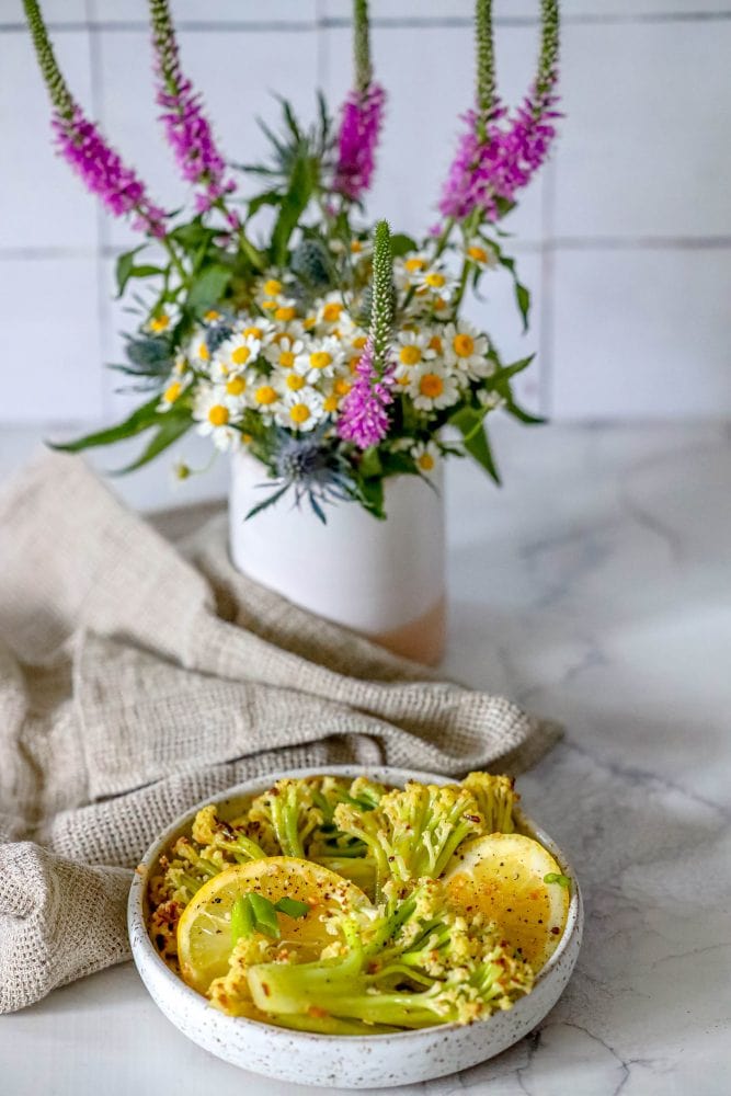 baby cauliflower and lemon slices in a white bowl on a marble countertop.  flowers in the background