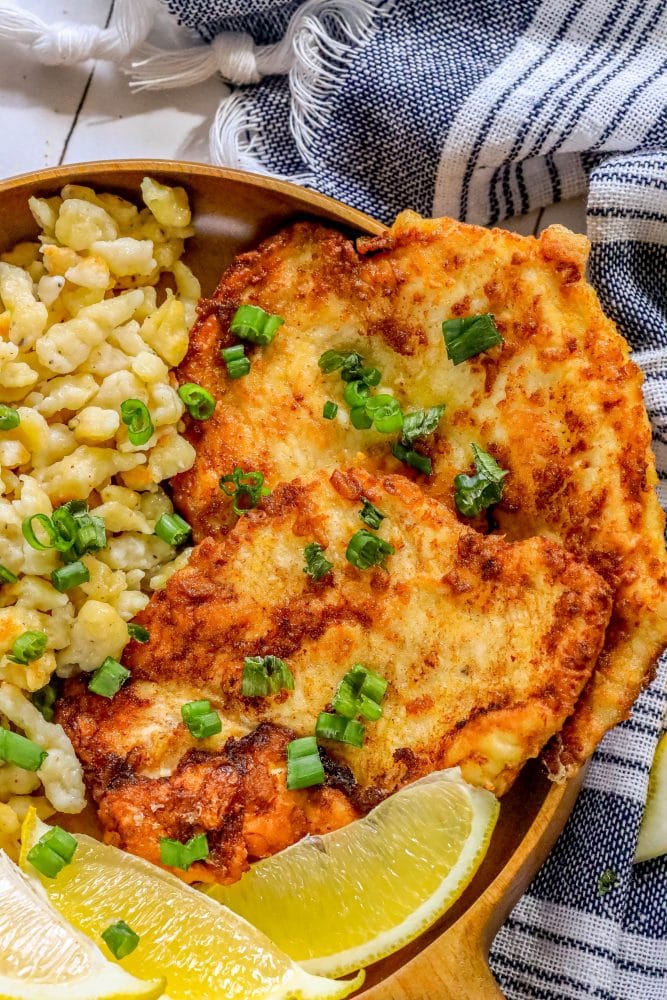 picture of breaded chicken schnitzel in a wooden bowl with green onions on top and lemon slices and spaetzle next to it