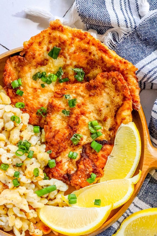 picture of breaded chicken schnitzel in a wooden bowl with green onions on top and lemon slices and spaetzle next to it