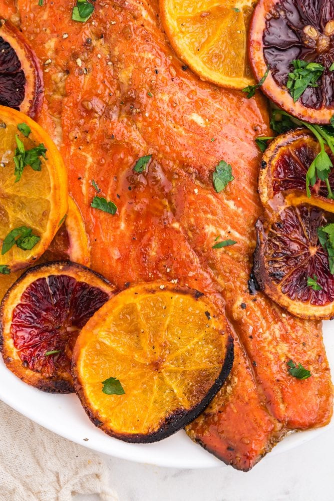 salmon with orange glaze on a white platter with browned oranges and chopped parsley on top on a table
