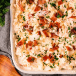A baking dish filled with creamy bacon chicken macaroni and cheese, topped with chopped bacon and parsley, placed on a gray cloth.