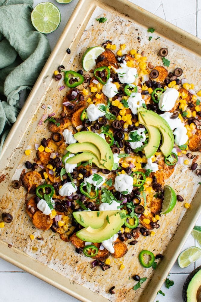 picture of sweet potato chip nachos on a plate with jalapenos, beans, corn, cheese, sour cream, avocados, and sliced limes