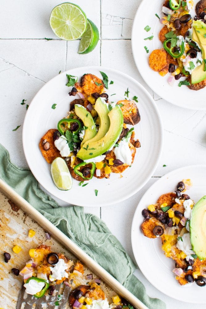 picture of sweet potato chip nachos on a plate with jalapenos, beans, corn, cheese, sour cream, avocados, and sliced limes