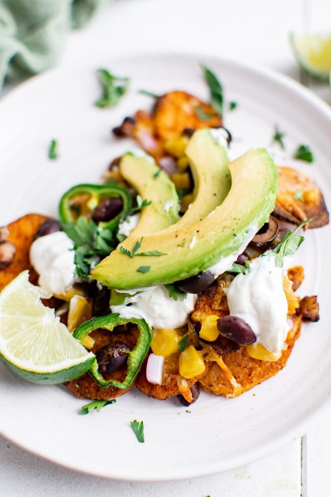 picture of sweet potato chip nachos on a plate with jalapenos, beans, corn, cheese, sour cream, avocados, and sliced limes