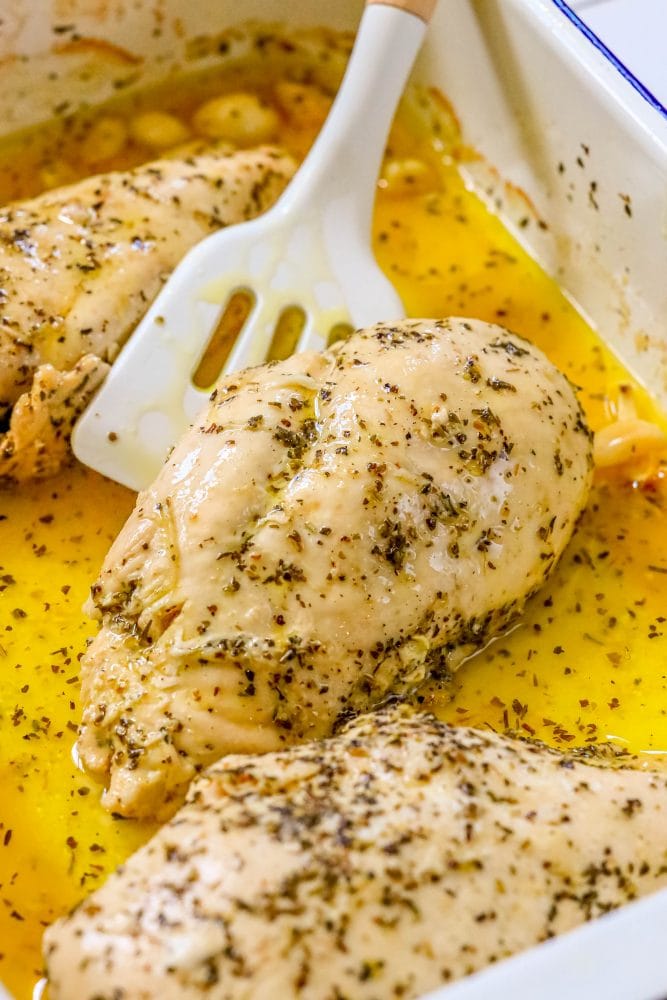chicken breasts in white baking dish being lifted by a white spatula, with melted butter, lemon juice, herbs, and garlic