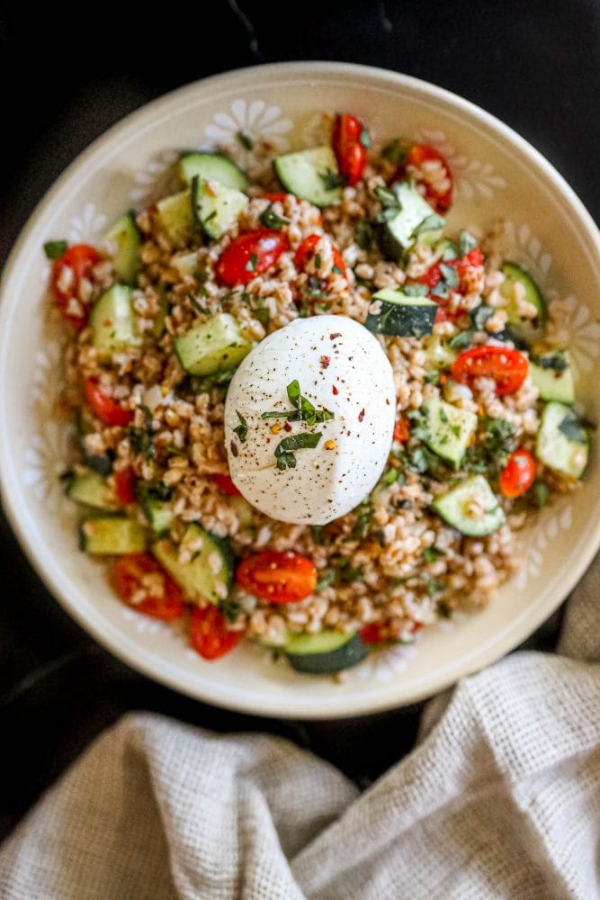 picture of farro salad with tomatoes, cucumber, basil in a bowl on a table with a burrata ball on top