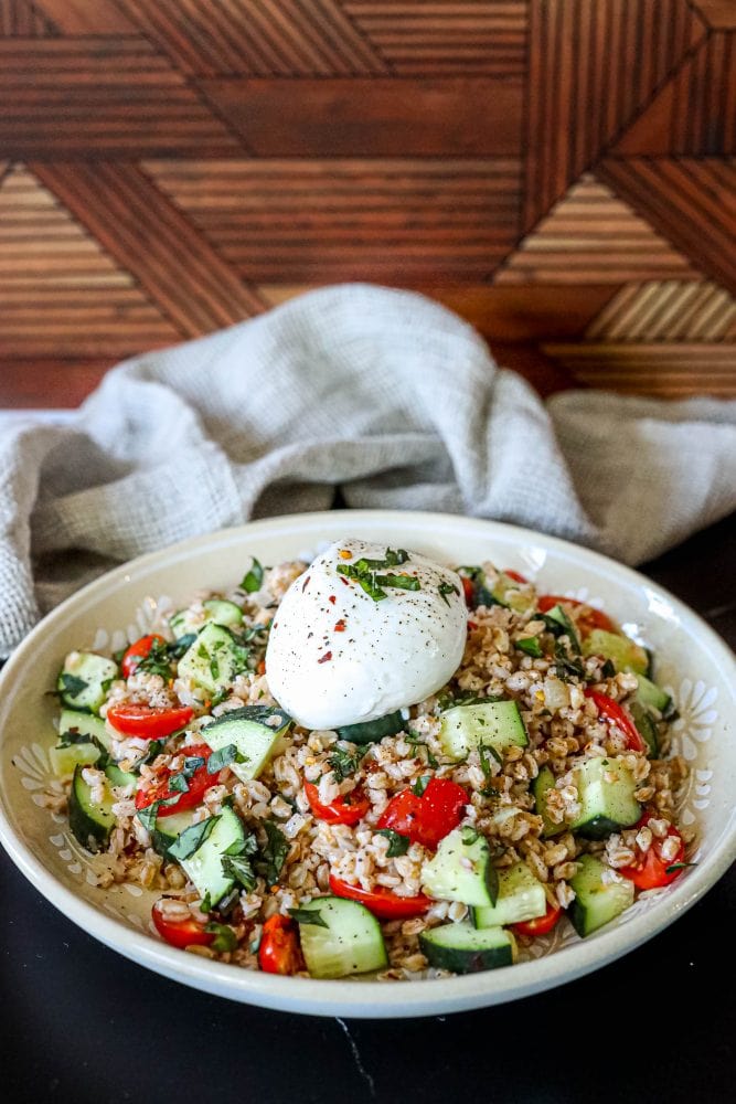 picture of farro salad with tomatoes, cucumber, basil in a bowl on a table with a burrata ball on top