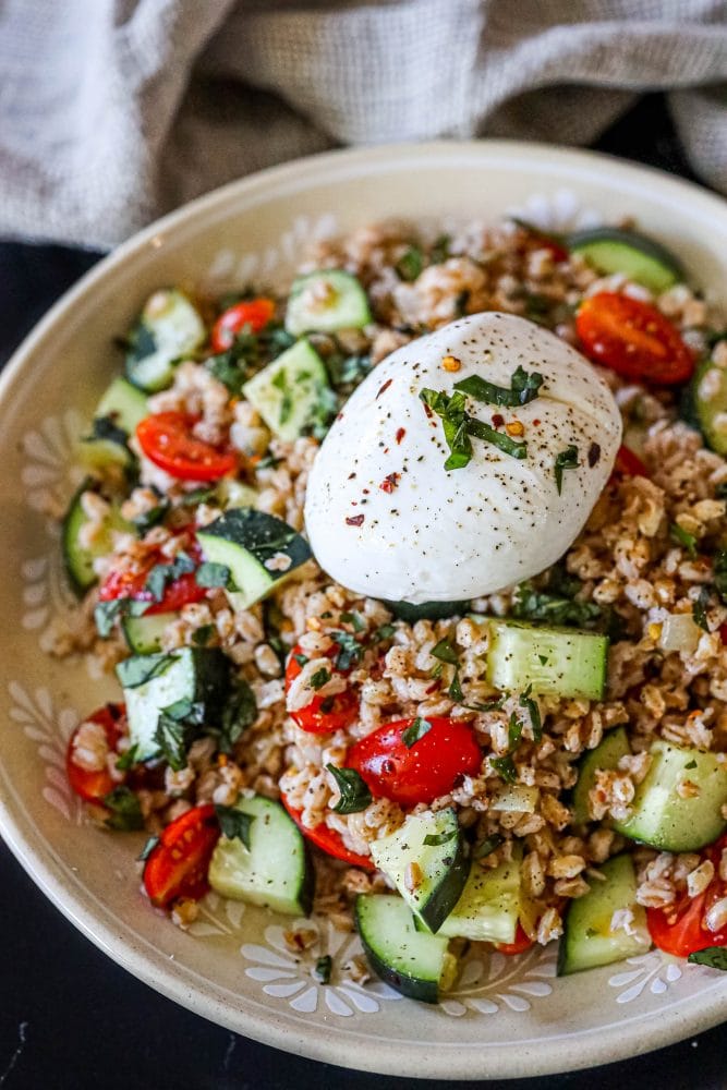 picture of farro salad with tomatoes, cucumber, basil in a bowl on a table with a burrata ball on top