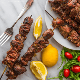 Two grilled Greek souvlaki skewers on a plate with lemon wedges, a fork, fresh arugula, and cherry tomatoes.
