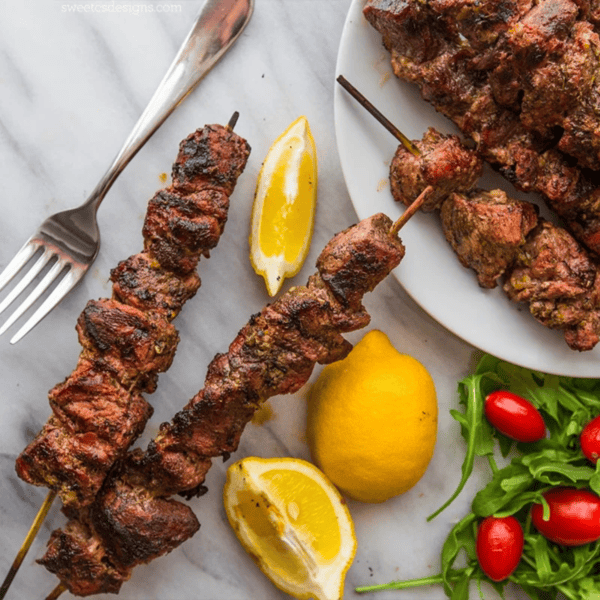 Two grilled Greek souvlaki skewers on a plate with lemon wedges, a fork, fresh arugula, and cherry tomatoes.
