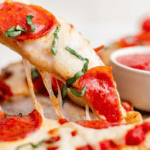 Close-up of a slice of pepperoni naan pizza with melted cheese and basil being lifted, with a bowl of tomato sauce in the background. Perfect for those seeking an easy keto pizza option.