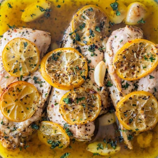 Cooked chicken breasts in a lemon and herb sauce, topped with lemon slices, chopped herbs, garlic cloves, and spices—all in one dish for The Best Easy Baked Garlic Lemon Chicken Breasts Recipe.