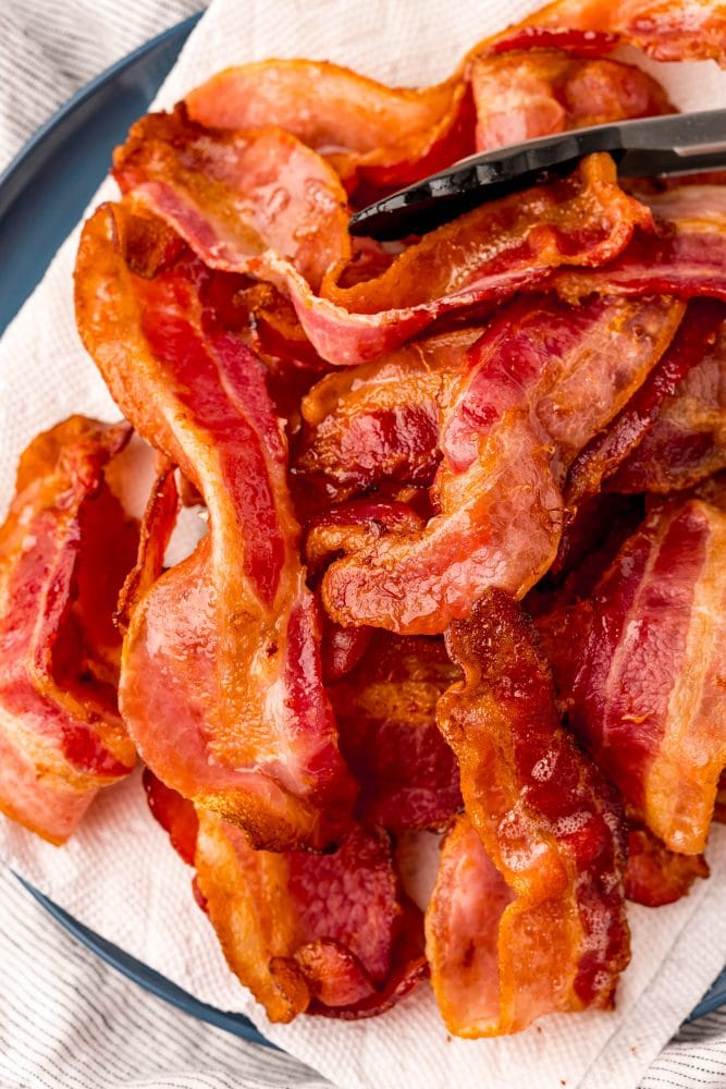 tongs lifting bacon onto plate with paper towels on it