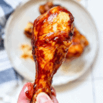 A hand holding an air fryer BBQ chicken drumstick gleams with a savory glaze, with a blurred plate of food in the background on a checkered tablecloth.
