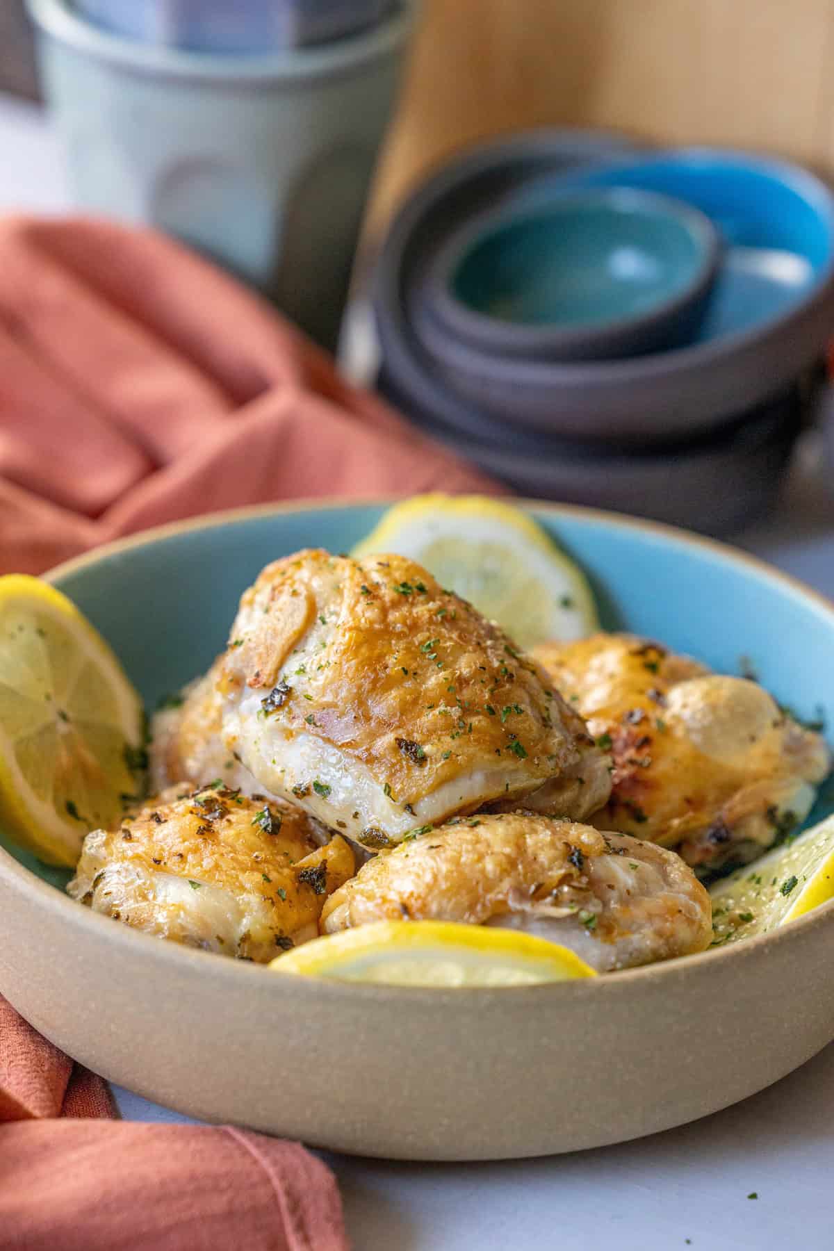 A bowl of easy air fryer lemon chicken thighs, garnished with lemon slices and herbs, sits enticingly in the foreground. Several stacked bowls and a cup linger in the background, setting the scene for this flavorful dish.