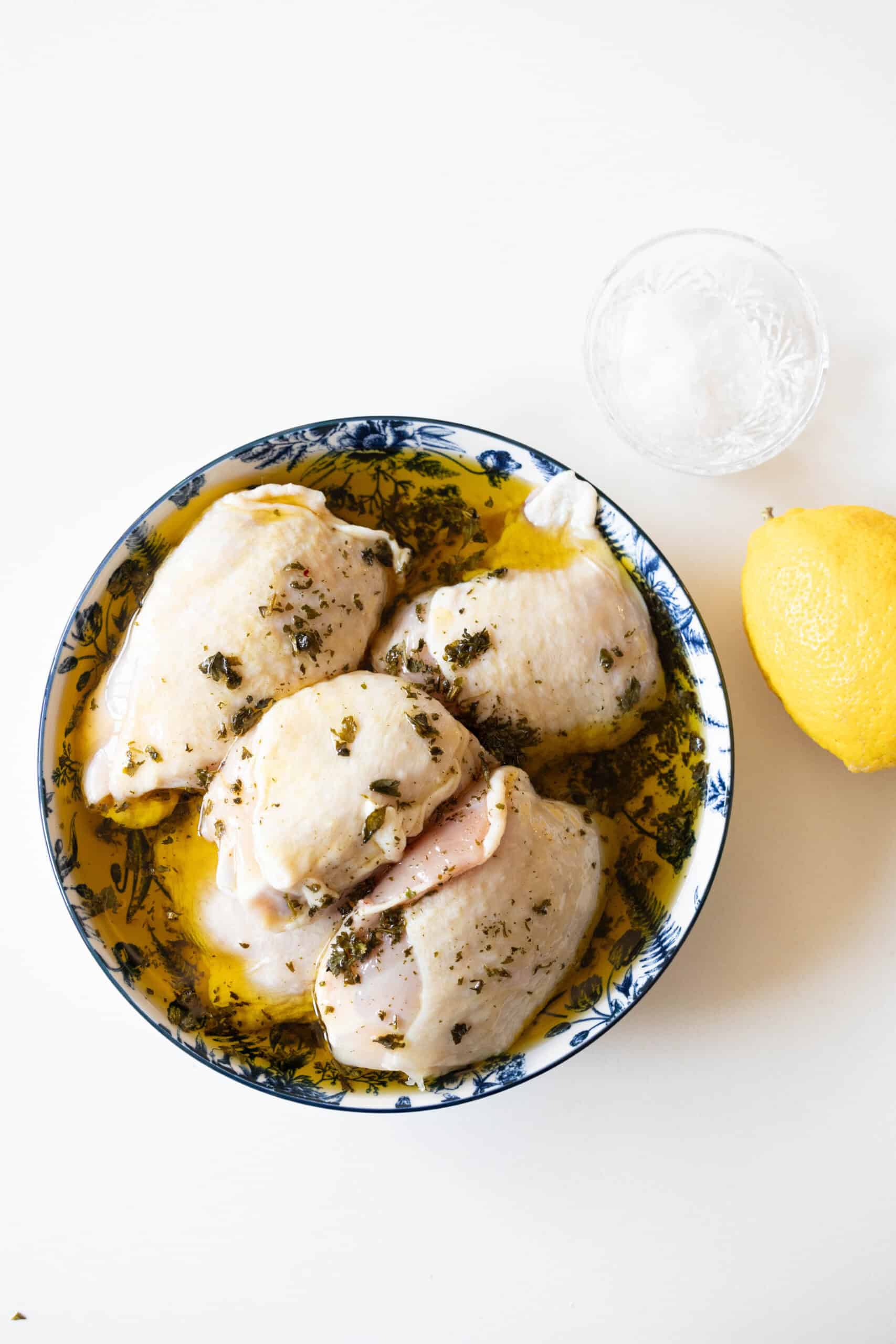 Raw chicken marinating in herbs and oil in a blue and white bowl, ready to become a delicious air fried lemon chicken. Nearby, a whole lemon and a small bowl of salt await their turn in this easy air fryer lemon chicken recipe.