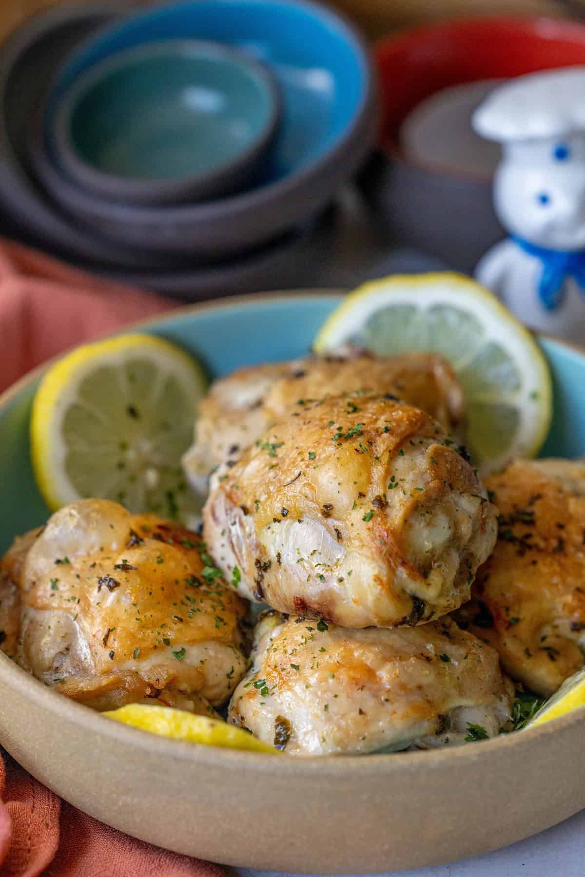 A bowl of seasoned air fried lemon chicken thighs garnished with parsley and lemon slices, with stacked bowls and a figurine in the background.