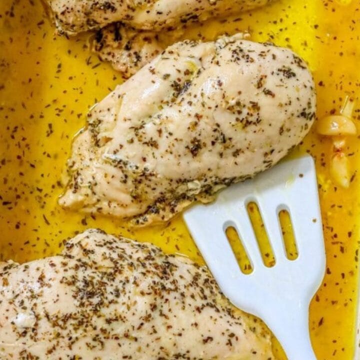 a chicken breast baked in melted butter, lemon juice, and garlic being lifted out of a casserole dish by a white spatula