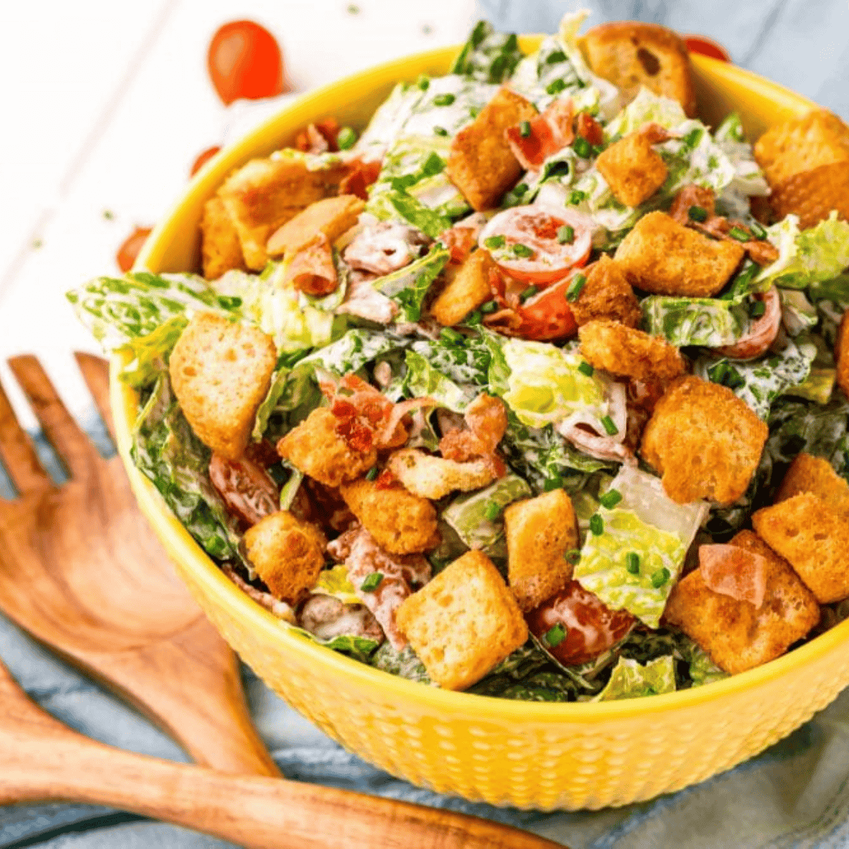 A yellow bowl filled with a BLT salad featuring croutons, cherry tomatoes, and bacon bits is placed on a blue cloth next to wooden salad utensils.
