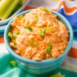 A blue bowl filled with creamy keto buffalo ranch chicken dip, garnished with chopped green onions, sits on a colorful striped cloth.