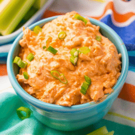 A blue bowl filled with creamy keto buffalo ranch chicken dip, garnished with chopped green onions, sits on a colorful striped cloth.