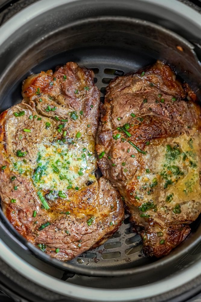 two steaks in an air fryer basket with butter and chives on them