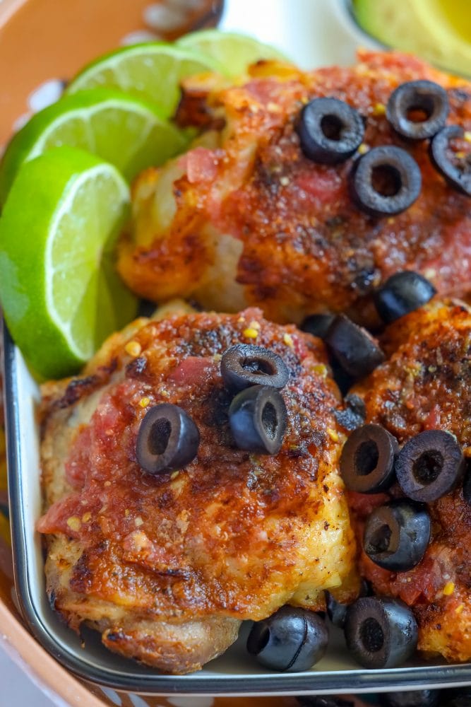 picture of chicken thighs toped with salsa and sliced black olives on a tray  with a bowl of salsa, sliced limes, and an avocado on the tray