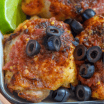 Close-up of air fried salsa chicken thighs topped with sliced black olives and tomato sauce, served with lime wedges.