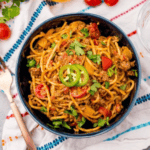 A bowl of spicy taco spaghetti with ground meat, sliced jalapeños, cherry tomatoes, and garnished with cilantro, viewed from above on a patterned cloth.