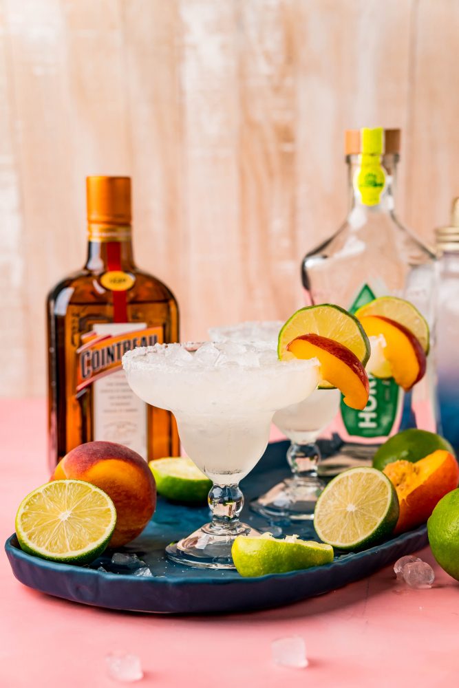 Picture of margarita glasses with peach slices in front of a bottle of tequila and a bottle of cointreau