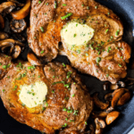Two cooked oven ribeye steaks topped with butter and chopped herbs, served in a skillet with sautéed mushrooms—a perfect example of the Best Oven Broiled Ribeye Steaks with Mushrooms.