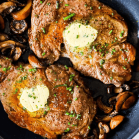 Two cooked oven ribeye steaks topped with butter and chopped herbs, served in a skillet with sautéed mushrooms—a perfect example of the Best Oven Broiled Ribeye Steaks with Mushrooms.