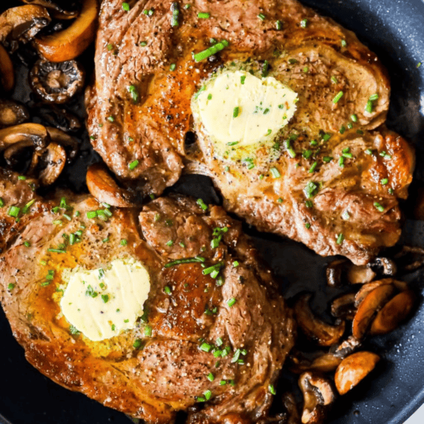 Two cooked oven ribeye steaks topped with butter and chopped herbs, served in a skillet with sautรฉed mushroomsโa perfect example of the Best Oven Broiled Ribeye Steaks with Mushrooms.