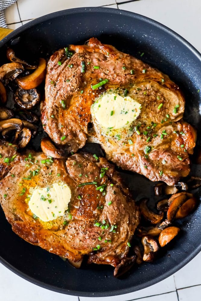 picture of ribeye steaks in a pan with mushrooms and pats of butter on top 