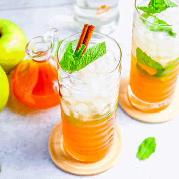 Two tall glasses of iced orange drink with mint leaves and cinnamon sticks, next to green apples, a glass jar of orange liquid, and a bottle—perfect inspiration for an Apple Cider Mojitos Cocktail recipe.