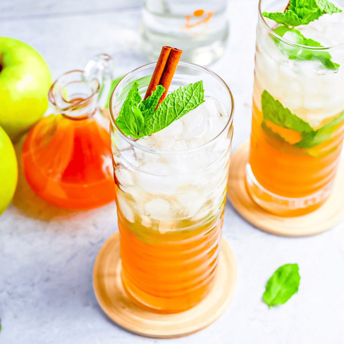 Two tall glasses of iced orange drink with mint leaves and cinnamon sticks, next to green apples, a glass jar of orange liquid, and a bottle—perfect inspiration for an Apple Cider Mojitos Cocktail recipe.