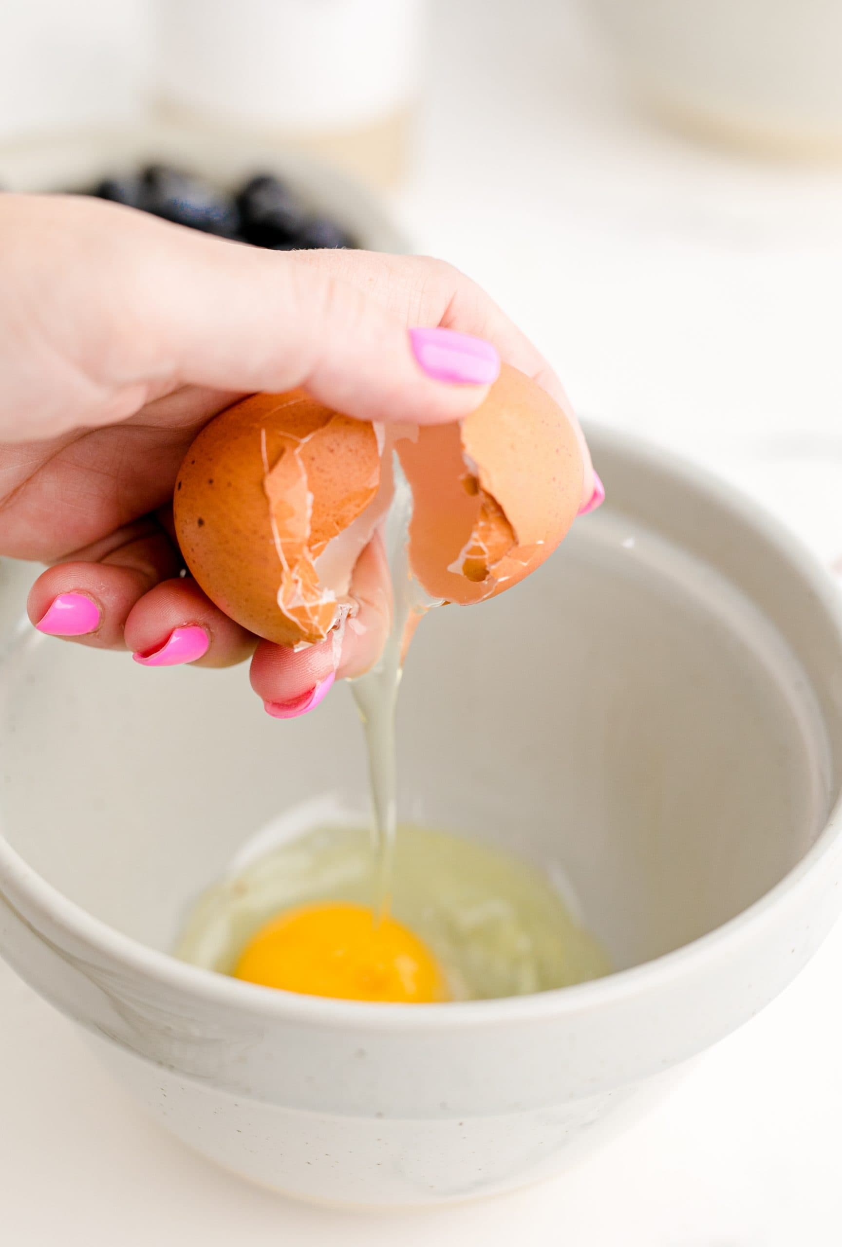 A person preparing a jumbo blueberry muffin recipe by breaking an egg into a bowl.