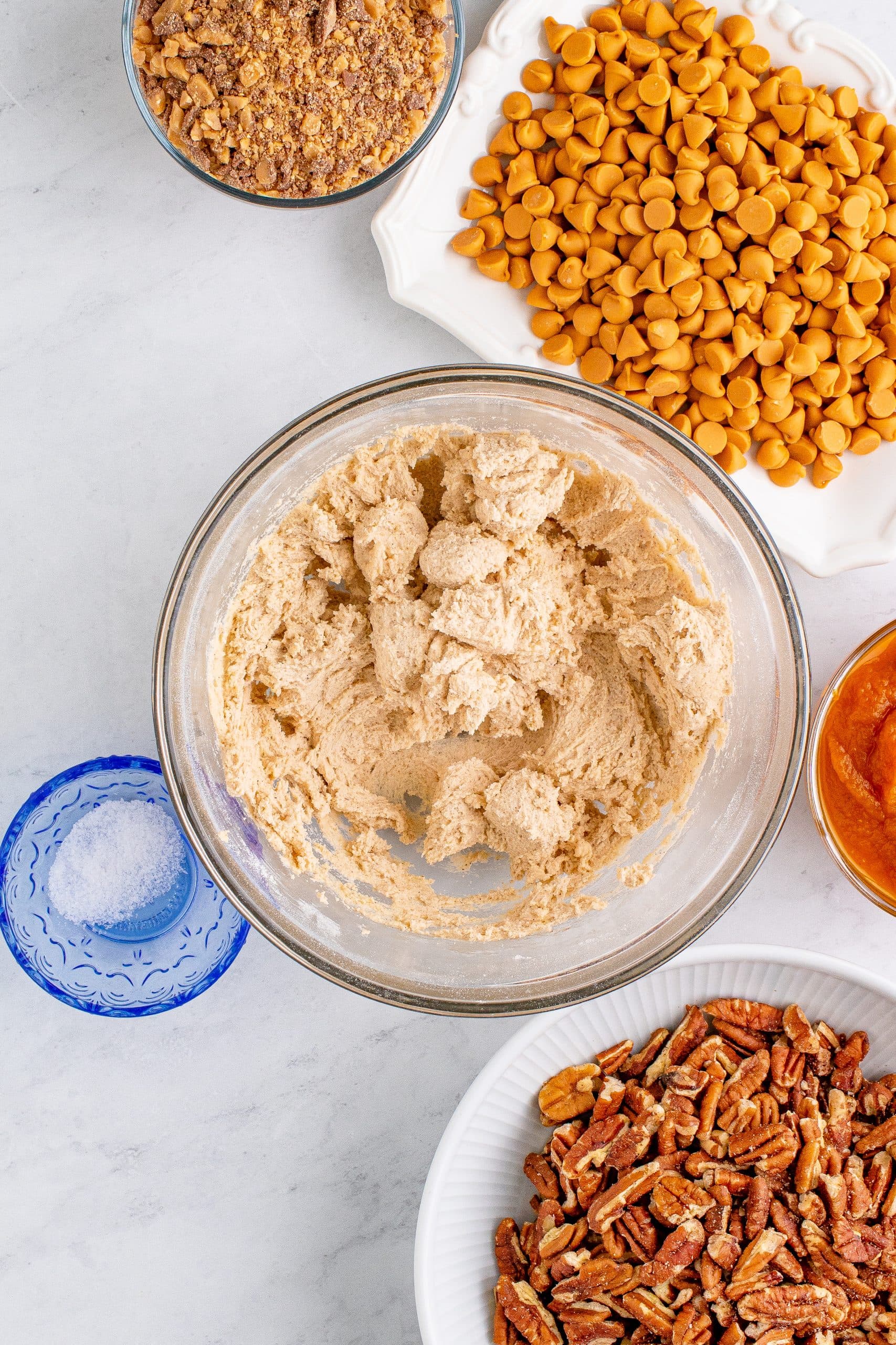 picture of cookie dough blended in a glass bowl next to butterscotch chips and nuts on a table
