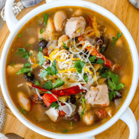 A bowl of spicy chicken soup with green chile and beans, topped with shredded cheese, chopped cilantro, and whole dried red chili peppers, placed on a wooden surface.