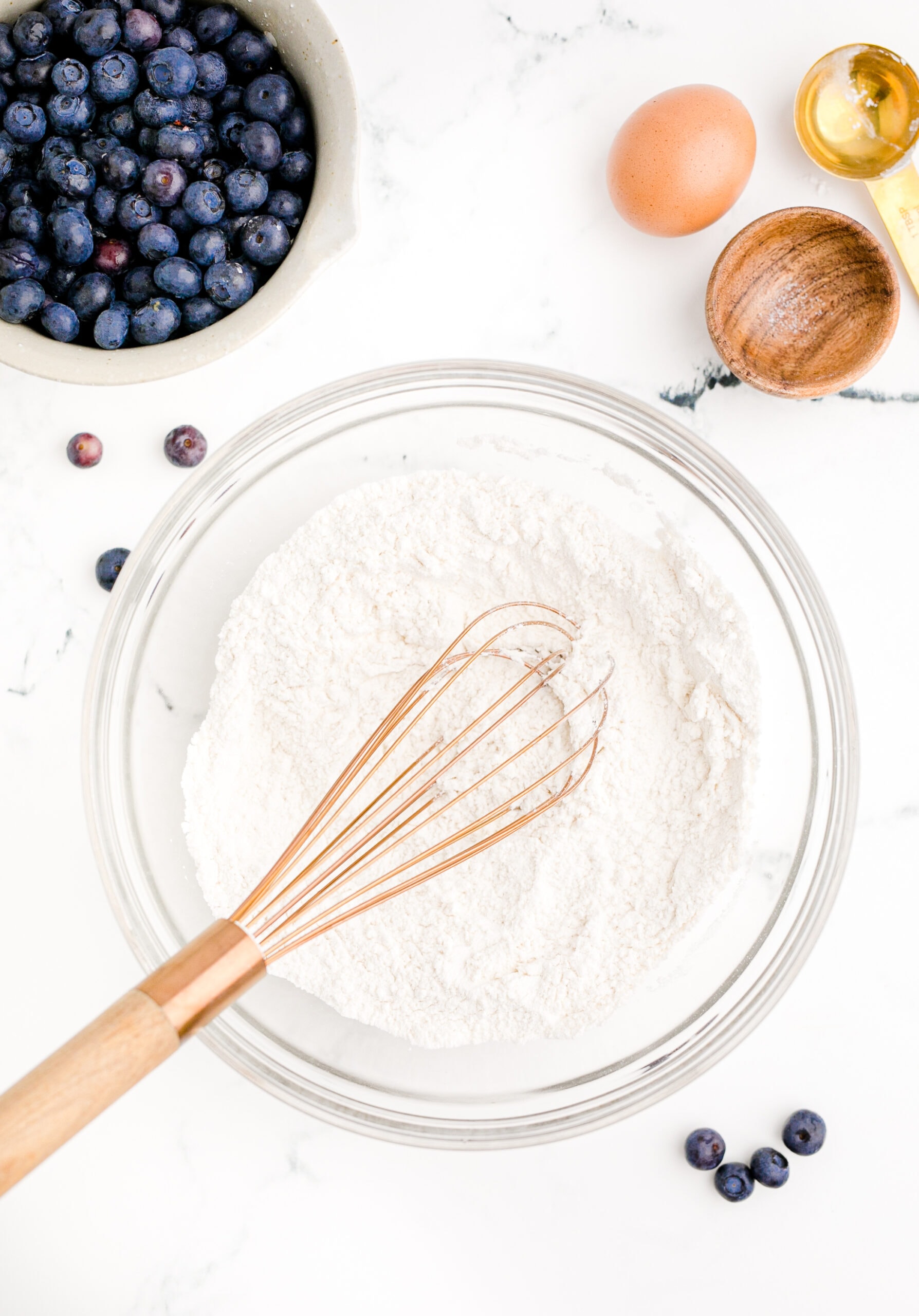 A bowl of flour, eggs, blueberries and a whisk.