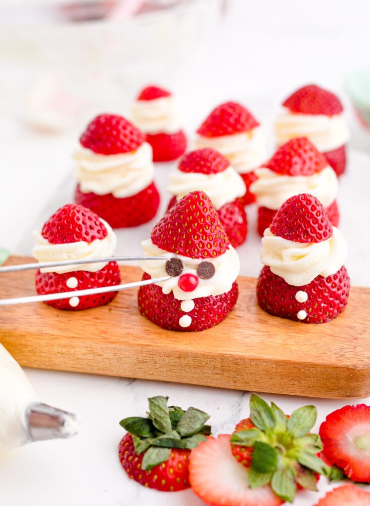 picture of tweezers putting chocolate chips on a strawberry sliced and topped with whipped cream and chocolate chips to look like santa