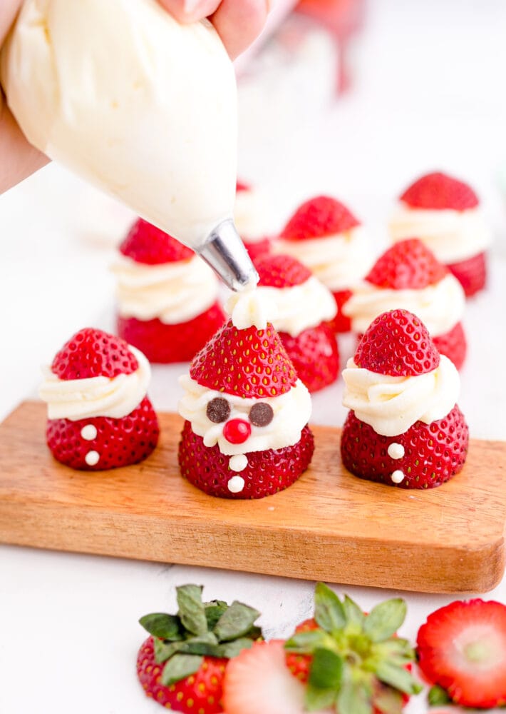 picture of whipped cream being piped onto a strawberry sliced and topped with whipped cream and chocolate chips to look like santa