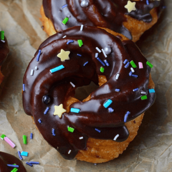 A close-up of a cruller donut covered in rich chocolate glaze and decorated with star-shaped, multicolored sprinkles. The cruller donut rests on brown parchment paper, highlighting its delicious detail.