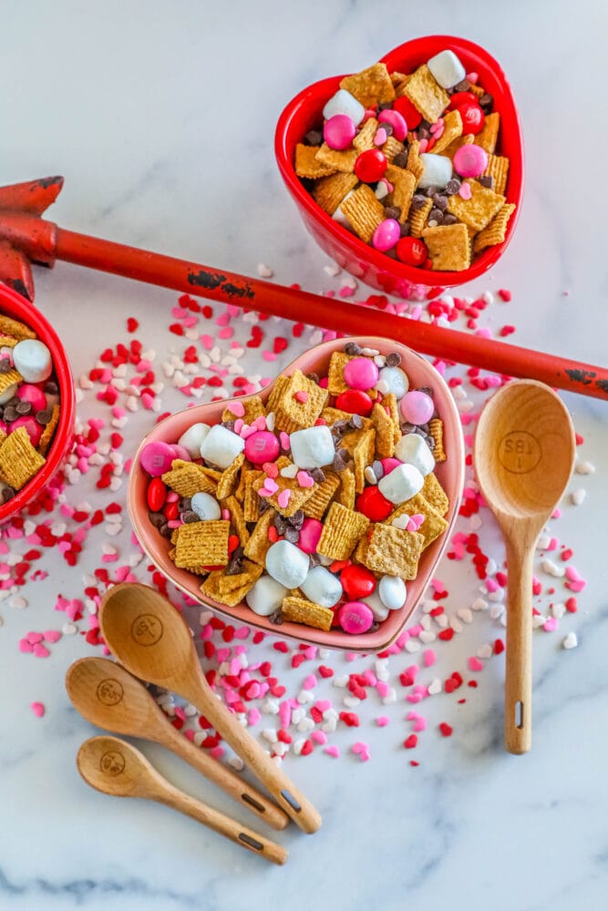 heart shaped bowl with snack mix made of marshmallows, pink m&ms, sprinkles,  and chocolate chips with wooden spoons around it
